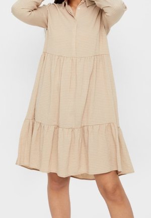 Femme portant une robe beige à manches longues, à volants, avec un col et une coupe ample, debout devant un fond uni clair.