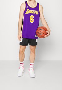 Maglia viola dei Lakers con dettagli gialli, pantaloni neri, scarpe bianche con dettagli rosa e una palla da basket.