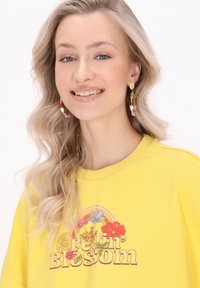 Žlutý svetr s grafikou "Feelin' Blossom", na které jsou květiny a duha. Modelka nosí barevné korálkové náušnice ve tvaru kruhu a má vlnité vlasy.