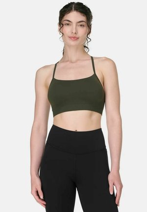 Vrouw draagt een donkergroene cropped top met dunne bandjes en zwarte leggings met hoge taille, staand tegen een effen witte achtergrond.