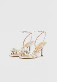 Silver strappy high-heeled sandals with a floral embellishment on the toe. Features a slim ankle strap and a shiny metallic finish.