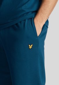 Tamsiai mėlynos šortai, pagaminti iš minkštos medžiagos, su auksiniu erelio logotipu priekyje ant kišenės. Elastinga juosmens juosta komfortui.