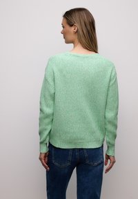 Femme aux longs cheveux bruns portant un pull en maille vert menthe et un jean bleu, debout face à un mur gris clair uni.