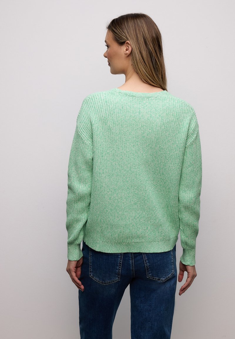 Femme aux longs cheveux bruns portant un pull en maille vert menthe et un jean bleu, debout face à un mur gris clair uni.