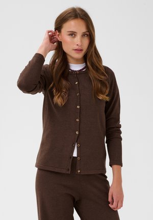 Femme aux longs cheveux bruns portant un cardigan marron boutonné sur un haut blanc et un pantalon marron assorti, debout devant un fond uni.