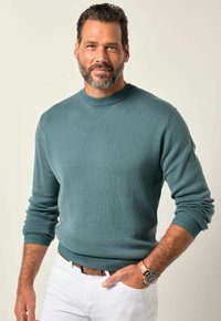 Suéter de cuello redondo en un suave tejido de punto en color verde azulado, con puños acanalados, combinado con pantalones blancos y un cinturón de cuero marrón. Acentuado con un reloj de pulsera.