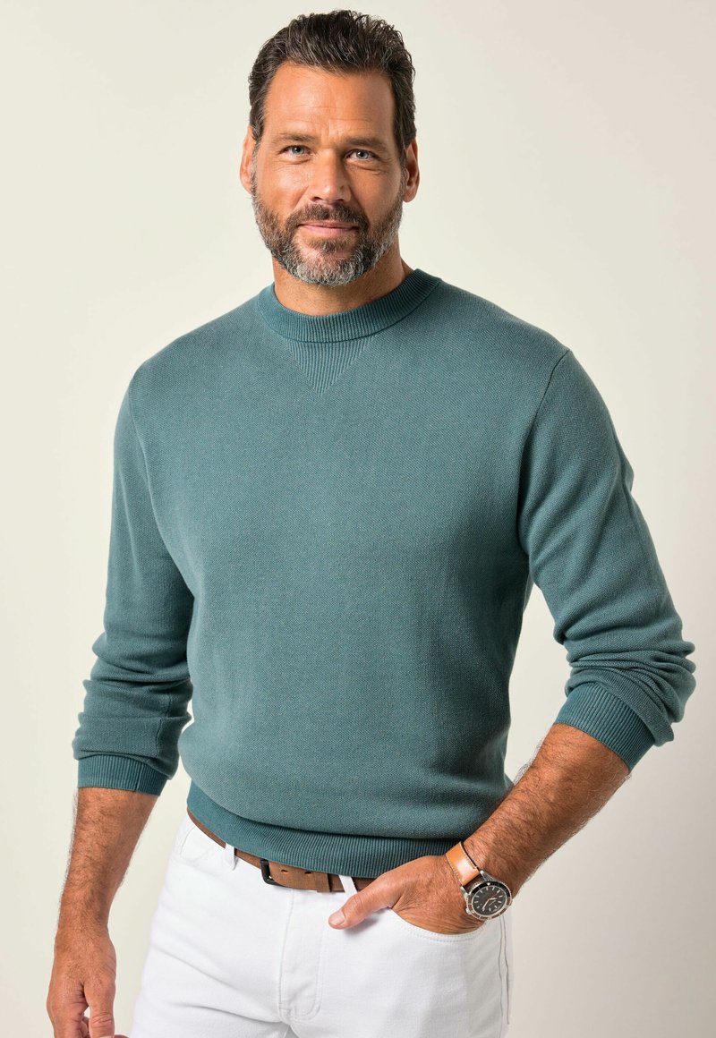 Suéter de cuello redondo en un suave tejido de punto en color verde azulado, con puños acanalados, combinado con pantalones blancos y un cinturón de cuero marrón. Acentuado con un reloj de pulsera.