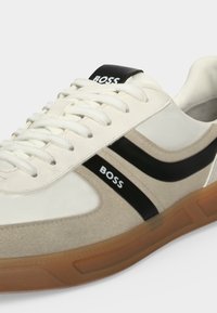 Baskets blanches et beiges avec des rayures noires et le texte "BOSS" sur le côté et la languette, avec des lacets blancs et une semelle en caoutchouc marron.