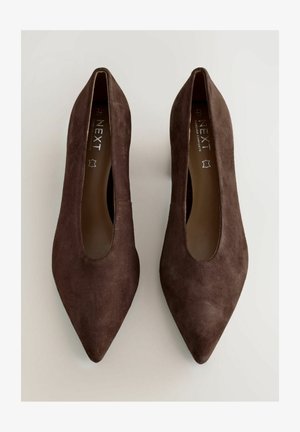 Brązowe zamszowe szpilki ze szpiczastym noskiem o smukłej sylwetce i otwartymi bokami. Wkładki mają wytłaczaną markę, co podkreśla ich wyjątkowy design.