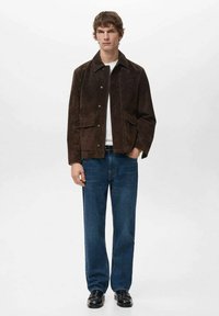 Chaqueta de ante marrón con cuello, cierres frontales de presión y dos grandes bolsillos tipo parche, combinada con vaqueros azules de pierna recta y zapatos negros.