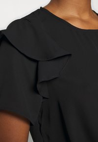 Schwarze Bluse mit kurzen Ärmeln und Rüscheldetails. Hergestellt aus leichtem Stoff mit glatter Textur und sauberem Nähen entlang der Kanten.
