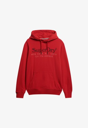 Sudadera con capucha roja con bolsillo tipo canguro en la parte delantera, capucha con cordón y logo "SuperDry" impreso. El material parece suave con una textura lisa.