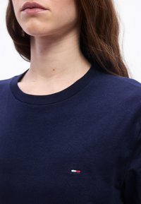 Γυναίκα που φοράει σκούρο μπλε μπλούζα με στρογγυλή λαιμόκοψη και μικρό κόκκινο, λευκό και μπλε λογότυπο στο στήθος, εμφανίζοντας το κάτω μέρος του προσώπου και τον λαιμό.