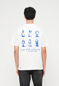 Άτομο που φοράει λευκό T-shirt με μπλε εικονογραφήσεις έξι μοντέρνων σχεδίων φωτιστικών και κείμενο "Φωτιστικά του 21ου Αιώνα 'Mid Century Modern'" στο πίσω μέρος.