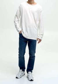 Homme portant une chemise henley blanche à manches longues, un jean bleu et des baskets blanches, debout avec une main dans la poche sur fond blanc.