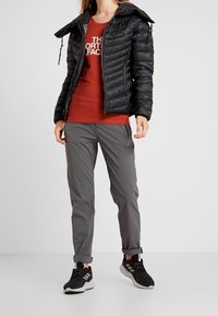 Svart puffjacka med grå insida, röd t-shirt, grå byxor och svarta sneakers; har dragkedjor och en viksöm på byxorna.