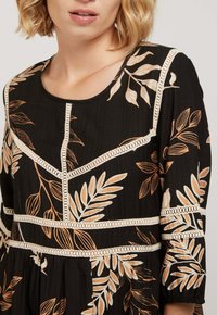 Robe noire avec un motif floral beige, dotée de dentelle et d'un col rond. Manches longues avec des poignets froncés et tissu texturé.