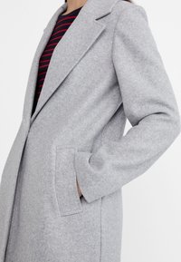 Lichtgrijze wollen jas met een gelapte kraag, diagonale zakken en een relaxte pasvorm. De stof heeft een zachte, gestructureerde uitstraling.