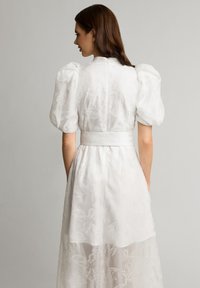 Biała sukienka z bufkami, kwiatowym wzorem i dopasowaną talią. Spódnica jest półprzezroczysta z warstwowym wzornictwem.