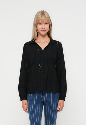 Svart langermet bluse med kragehals, med et teksturert mønster og utskjæringsdetaljer. Brukt med stripete jeans.