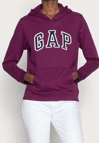 Felpa con cappuccio color bordeaux con la scritta "GAP" in bianco e blu navy, tasca frontale e polsini a costine, abbinata a pantaloni bianchi.