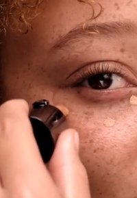 Applicateur de fond de teint utilisé sur une peau avec des taches de rousseur ; des taches beige chaud visibles, soulignant la texture liquide et bien mélangée du produit.