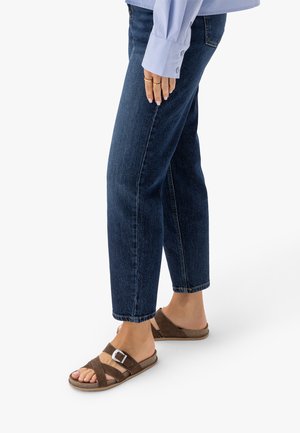 Osoba ubrana w ciemnoniebieskie dżinsy, jasnoniebieską koszulę z długim rękawem oraz brązowe zamszowe sandały z klamrami, stojąca na białym tle.