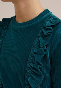 Zelena rebrasta pulover s okroglim izrezom, ki ima naborke ob straneh. Mehka tekstura poudarja igriv oblikovalski detajl.