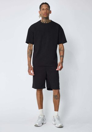 Homme avec des tatouages portant un T-shirt noir oversize, un short noir, des chaussettes blanches et des baskets blanches, debout devant un fond clair uni.