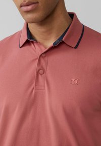 Roza polo majica z mornarsko barvo ovratnika, s poudarkom na dveh gumbih in teksturirano tkanino. Majhna, vtisnjena oznaka je nameščena na prsih.