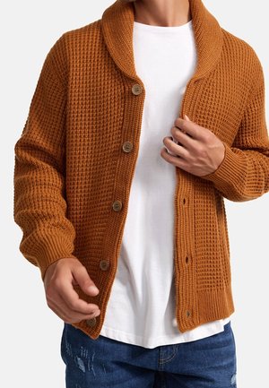 Homme portant un cardigan en maille gaufrée couleur rouille sur un t-shirt blanc et un jean bleu déchiré, en train de fermer le bouton du milieu.