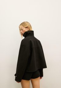 Giacca nera oversize con collo alto e cuciture a vista, realizzata in tessuto strutturato. Il design presenta maniche larghe e bottoni ai polsini.