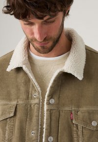 Veste en velours côtelé vert olive avec col en shearling blanc, boutons en métal argenté et un petit logo Levi's rouge sur la poche poitrine.