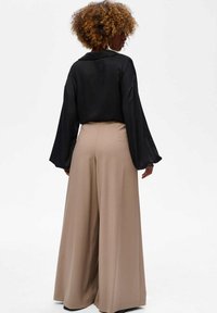 Zijdezwarte blouse met brede mouwen, gecombineerd met hooggerokte, beige wijde broekspijpen. Soepele texturen, nauwkeurige snit en een neutraal kleurenpalet.