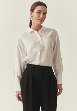Biela košeľa na zapínanie s textúrovaným vzorom, dlhými rukávmi a klasickým golierom, kombinovaná s čiernymi nohavicami s vysokým pásom.