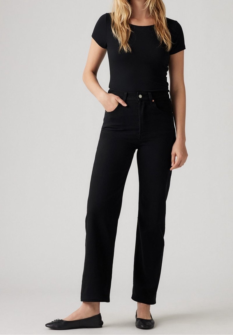 Femme portant un haut noir à manches courtes, un jean noir taille haute et des ballerines noires, debout avec une main dans la poche devant un fond uni.