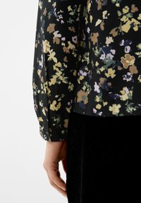 Schwarze Bluse mit floralem Muster und kurzen, umgeschlagenen Ärmeln; zeigt verschiedene Grüntöne, Gelb- und Lavendel-Muster. Weiches, strukturiertes Material.