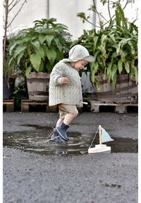 Egy világoszöld steppelt kabátot és sapkát viselő gyerek pocsolyában játszik, tengerészkék gumicsizmában. Egy kis vitorláshajó világoskék vitorlával a közelben.