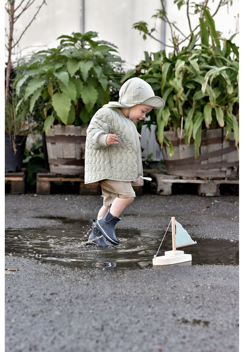 Barn i en ljusgrön vadderad jacka och hatt leker i en vattenpöl, bär marinblå gummistövlar. En liten segelbåt med ljusblå segel i närheten.