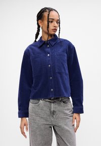 Camicia di velluto a coste blu navy con colletto, bottoni frontali e due tasche sul petto. Maniche lunghe con tessuto strutturato. Indossa jeans grigi.