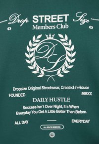 Grünes Baumwoll-T-Shirt mit weißem Drucktext. Es verfügt über ein kreisförmiges Logo mit einer Krone, Lorbeerblättern und verschiedenen motivierenden Sprüchen.