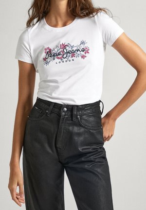 Frau trägt ein weißes Pepe Jeans London T-Shirt mit pinkem Blumenmuster und hoch taillierte schwarze Lederhose, Hand auf der Hüfte.