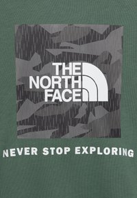 T-shirt in cotone verde con un quadrato in camouflage grigio con il logo "THE NORTH FACE" in bianco e il testo "NEVER STOP EXPLORING" sotto.