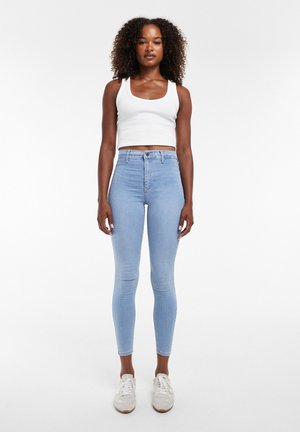 Kvinde stående iført hvid cropped tanktop, højtaljede lyseblå skinny jeans og hvide sneakers mod ensfarvet baggrund.