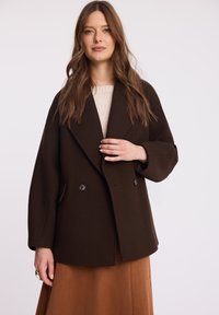 Podwójnie zapinany brązowy wełniany płaszcz z szerokimi klapami i dużymi czarnymi guzikami. Posiada luźny krój oraz boczne kieszenie.