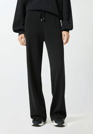 Pantaloni neri a gamba larga realizzati in un tessuto morbido, con vita regolabile tramite coulisse e cuciture minime. Abbinati a scarpe da ginnastica nere.