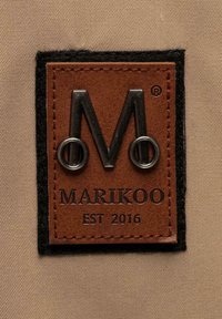 Étiquette en cuir marron avec une bordure en feutre noir, présentant une lettre "M" en argent en relief et le texte "MARIKOO EST 2016" embossé en dessous.