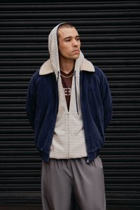 Marineblå kordfløjs zip-jakke med krave af cremefarvet faux fur, lagdelt over en beige stribet hættetrøje og grå bukser.