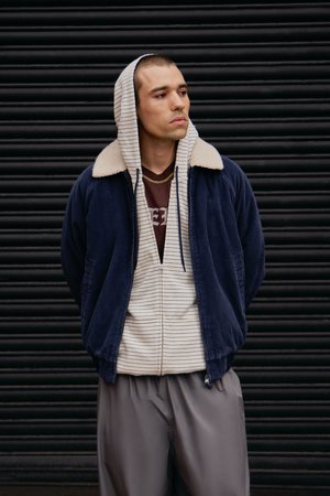 Veste zippée en velours côtelé bleu marine avec col en fausse fourrure crème, superposée sur un sweat à capuche beige à rayures et un pantalon gris.