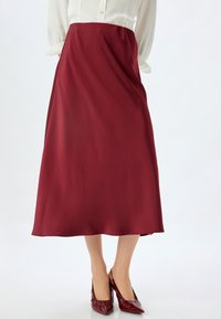 Maroon satin A-linjainen hame, keskipitkä, sileä tekstuuri, yhdistetty kiiltäviin purppuranpunaiseen korkokenkiin, esitetty neutraalilla taustalla.